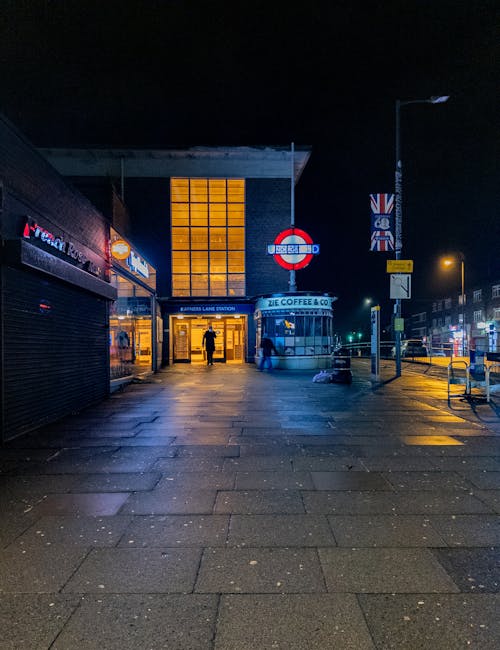 A nighttime scene outside a transport station with illuminated entrance doors, where a person is standing near the doorway. The surrounding pavement is wet and reflects the yellow streetlights. To the right, a bus stop sign and a lamppost are visible, along with a small kiosk advertising ZIE Coffee & Co. and a UK flag on a pole. In front of the station, there are parked boxes and luggage, indicating a home relocation or moving process. The environment is quiet and well-lit, with an urban setting and buildings lining the street. This scene reflects the logistics of furniture transport and packing during a house move, with [COMPANY_NAME], such as Man with Van Arnos Grove, likely providing related removals services in this area.