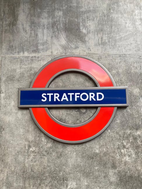 A close-up photograph of a circular London Underground station sign featuring a red ring and a blue horizontal bar with the word 'STRATFORD' in white capital letters. The sign is mounted on a textured grey concrete wall, indicating a station entrance or interior. This image relates to the context of home relocation or moving services provided by Man with Van Arnos Grove, with the sign representing a key transportation hub near Arnos Grove tube station, relevant for furniture transport and logistics during moving and packing processes.