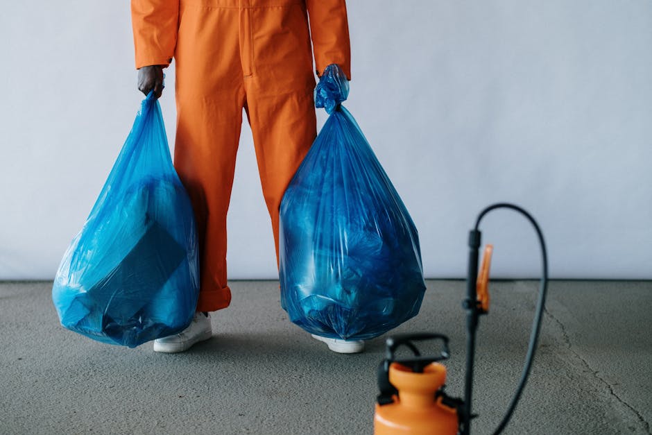 A person dressed in orange work overalls and white shoes stands indoors on a concrete floor, holding two large blue plastic rubbish bags filled with waste. The individual’s face is not visible, only their body from the waist down. In the background, a plain light-colored wall is visible, indicating a clean, indoor environment typical of a home or storage space prepared for packing or rubbish collection during a house removal or relocation process. On the right side of the image, part of a black and orange sprayer or cleaning device is visible, suggesting cleaning or waste management activity related to packing or clearing out a property. The scene reflects a packing and moving or waste removal task as part of home relocation services often handled by companies like Man with Van Arnos Grove, supporting efficient furniture transport, packing, and loading processes involved in house removals.