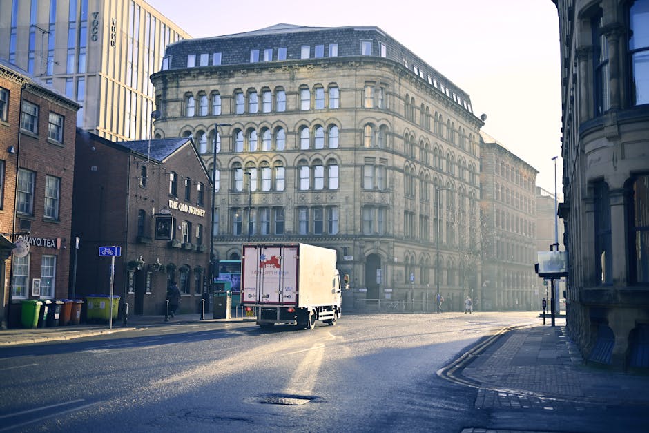 A street scene showing a row of multi-storey residential buildings with balconies and large windows next to a pavement on a clear day. Parked cars line the roadside, and pedestrians are walking along 