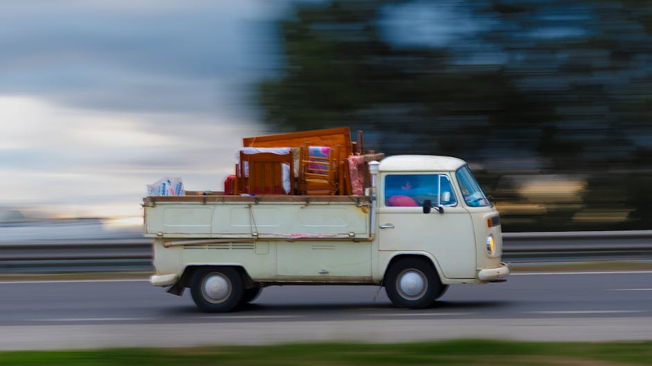 A vintage cream-colored van with a compact, rounded cab and large front windshield is shown driving on a road with a blurred background indicating motion. The van's flatbed cargo area is loaded with v