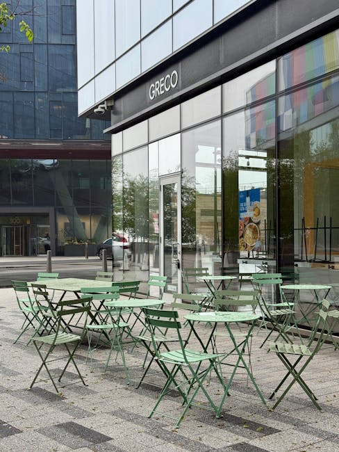 An outdoor area in front of a modern glass-fronted building with the sign 'GRECO' visible above the entrance. Several light green metal tables and matching chairs are arranged on a paved surface, with some people sitting at a few tables. The building's large glass windows reflect nearby trees, parked cars, and the surrounding urban environment. The scene is well-lit with natural daylight, and the setting appears to be part of a restaurant or café located within a commercial complex. This exterior space could serve as a waiting or outdoor dining area, commonly associated with restaurants offering convenient access for customers during house removals or moving activities to ensure smooth logistics and loading processes.