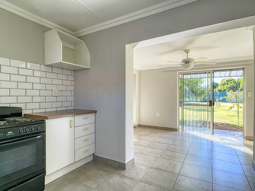 Interior view of a residential property showing a kitchen area with a black freestanding oven and stove, white cabinetry with light-colored handles, and a pinkish countertop. Above the countertop, there is a white tiled backsplash with a brick pattern and an open wall-mounted cabinet. Adjacent to the kitchen, the image extends to a bright, unfurnished living space with tiled flooring, cream-colored walls, a ceiling fan, and a large sliding glass door leading to an outdoor garden area with greenery and trees. The scene depicts an ongoing home relocation process, with the space prepared for packing and furniture transport by a professional removals service such as Man with Van Arnos Grove.