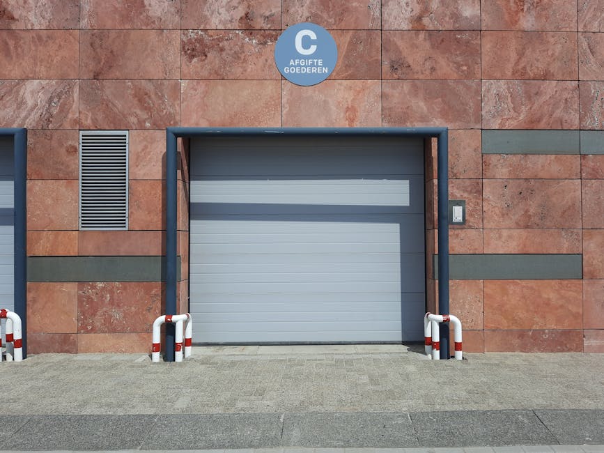 A grey, fully closed commercial roller shutter door set within a brick and stone exterior wall of a building, with a small illuminated intercom panel to the right side. The area in front of the shutte