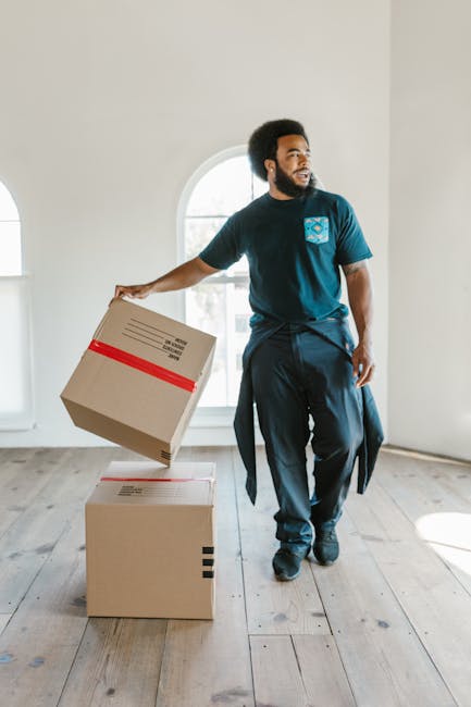 A man with dark, curly hair and a beard, dressed in a navy blue t-shirt with a small logo on the chest and dark trousers, is inside a bright, empty room with white walls and wooden flooring. He is holding a cardboard moving box with red and black tape, preparing to lift or move it during a home relocation process. Behind him, there is a large arched window letting in natural light, casting shadows across the floor. The room appears to be part of a house or flat awaiting furniture and packing, with additional boxes stacked on the floor nearby. The scene reflects the loading and packing stage of house removals, likely handled by a professional service such as Man with Van Arnos Grove, as part of their furniture transport and moving logistics. The environment is tidy and uncluttered, emphasizing a careful and organized move.
