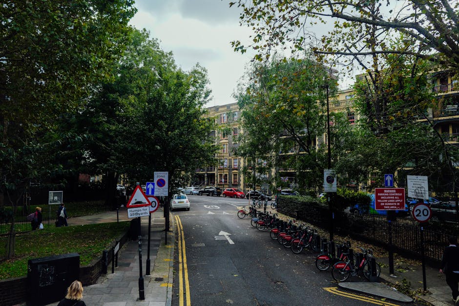 A view of a residential street with several parked bicycles secured to a bike rack on the right side, and various traffic and parking signs, including a speed limit of 20 miles per hour, a no parking zone, and a warning sign about humped crossings. The street is lined with tall trees with green and yellow leaves, suggesting an autumn setting, and bordered by multi-storey buildings with balconies and large windows. On the left, there is a sidewalk with a few pedestrians walking, one carrying a bag, and a small fenced green space with grass and shrubs. The road surface appears dark and slightly wet, with yellow line markings along the edge. The overall scene depicts an urban environment suitable for house removals, with a focus on the logistical considerations of moving furniture and boxes through an area with posted parking and traffic regulations, hinting at the challenges and planning involved in home relocation services like those offered by Man with Van Arnos Grove.