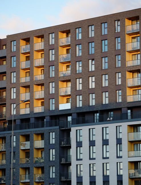 Exterior view of a modern high-rise building with multiple floors, featuring a combination of dark grey, light grey, and yellow painted sections. The building has several glass balconies with transparent railings and multiple evenly spaced windows, some of which are open. The lower part of the building includes a black metal fence and entrance area. The image shows a clear sky with some clouds, suggesting daylight, and is associated with house removals and home relocation services offered by Man with Van Arnos Grove, highlighting challenges and logistics of moving into high-rise flats on Green Lanes.