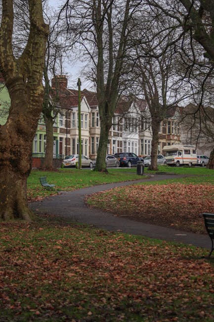 Photograph of a residential street near Grovelands Park in Arnos Grove, showing a curved pathway within a small park area with fallen autumn leaves scattered on the ground. Two benches are visible, po